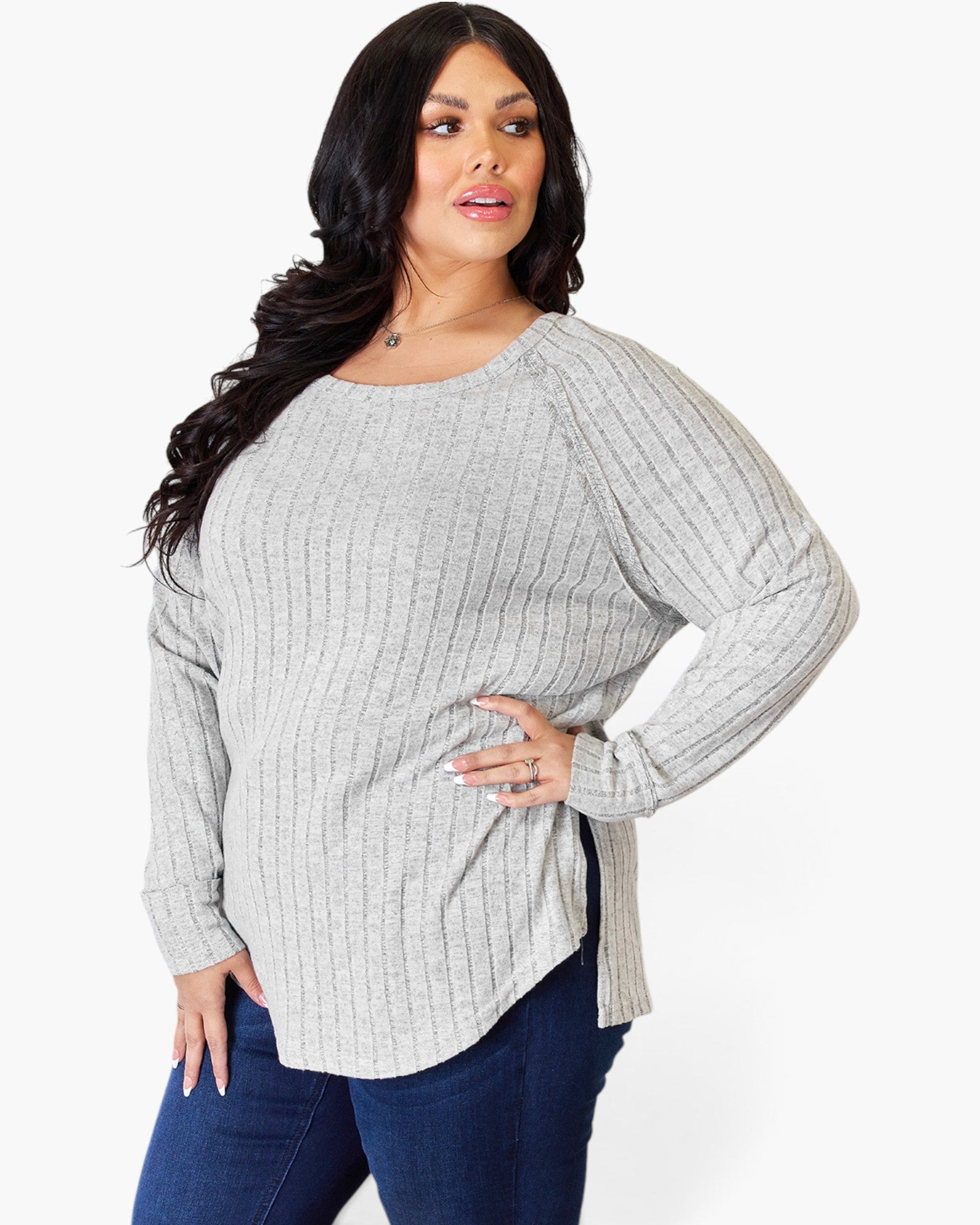 A woman modeling a light gray ribbed knit long-sleeve top with a boat neckline and side slit, paired with dark blue jeans against a plain white background.