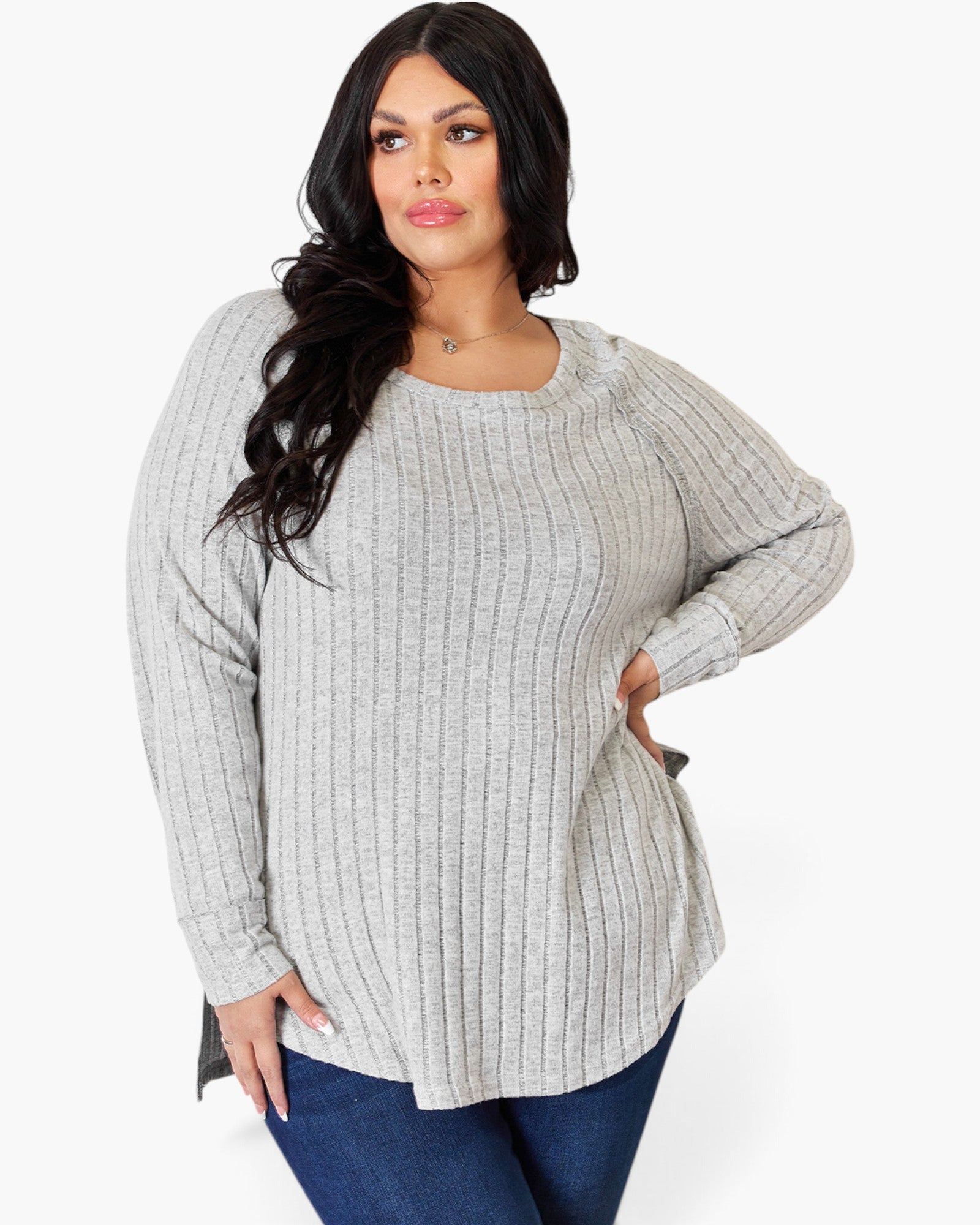 A woman modeling a light gray ribbed knit sweater with long sleeves and a crew neckline, paired with dark blue jeans, against a plain white studio background.