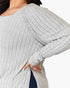 A close-up of a woman wearing a light gray ribbed knit top with long sleeves and a side slit, with one hand resting on the hem.