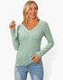A woman wearing a sage green ribbed knit long-sleeve V-neck sweater and light blue jeans, posing against a solid white studio background.