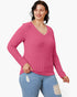 A woman wearing a pink ribbed long-sleeve V-neck sweater and light blue distressed jeans, standing against a plain white studio background.