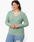 A woman wearing a sage green ribbed knit long-sleeve V-neck sweater and light blue jeans, standing against a plain white studio background.