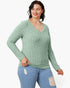 A woman modeling a light sage green ribbed knit long-sleeve V-neck sweater with light blue distressed jeans against a plain white background.