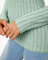 A close-up of a person wearing a ribbed knit sweater in a light sage green color, paired with light blue jeans, against a plain white background.