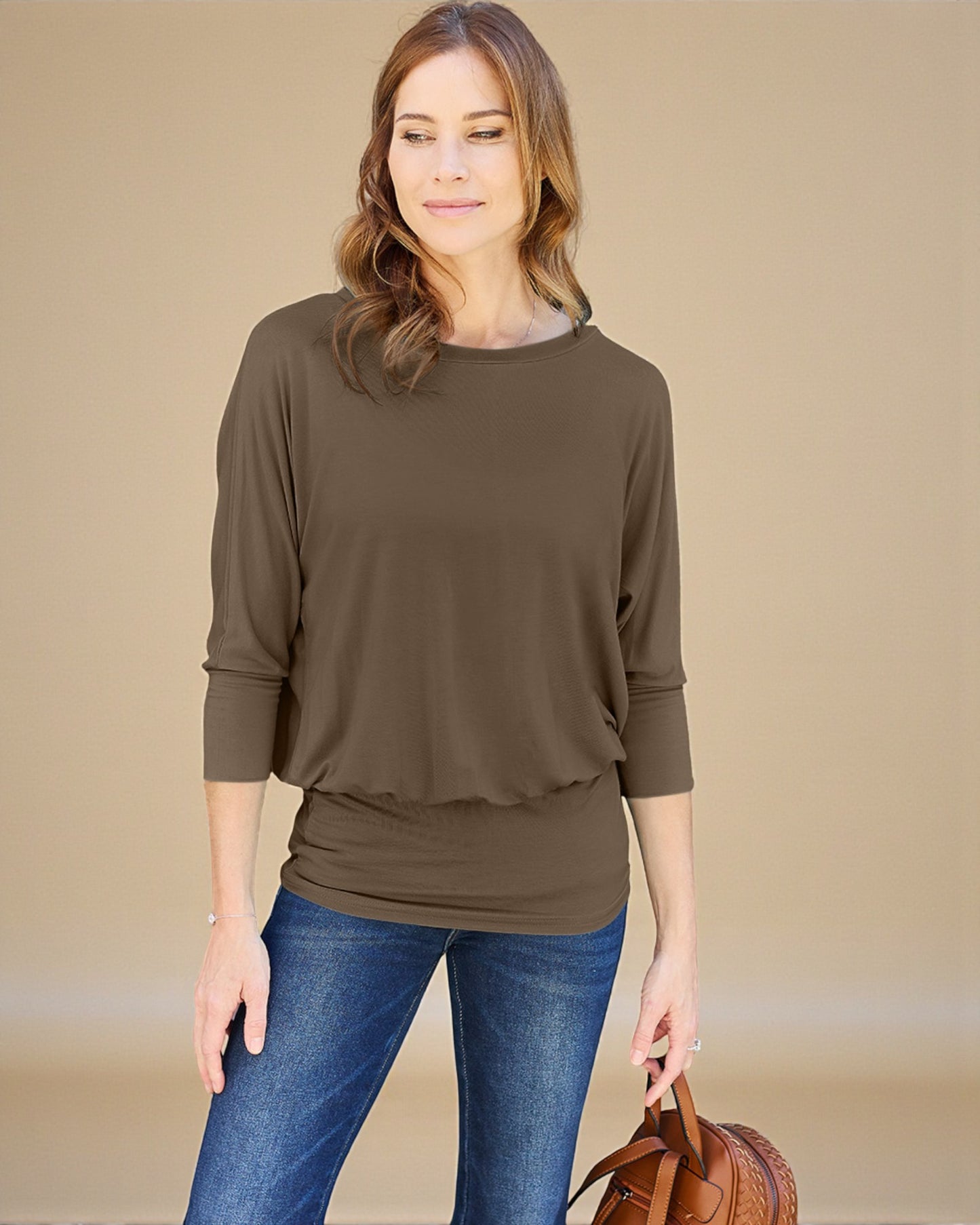 Woman wearing a brown long-sleeve top and blue jeans, holding a brown leather bag on a beige background