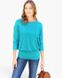 A woman wearing a bright turquoise three-quarter sleeve top with a ruffled hem, paired with blue jeans, holding a brown leather handbag against a plain white background.