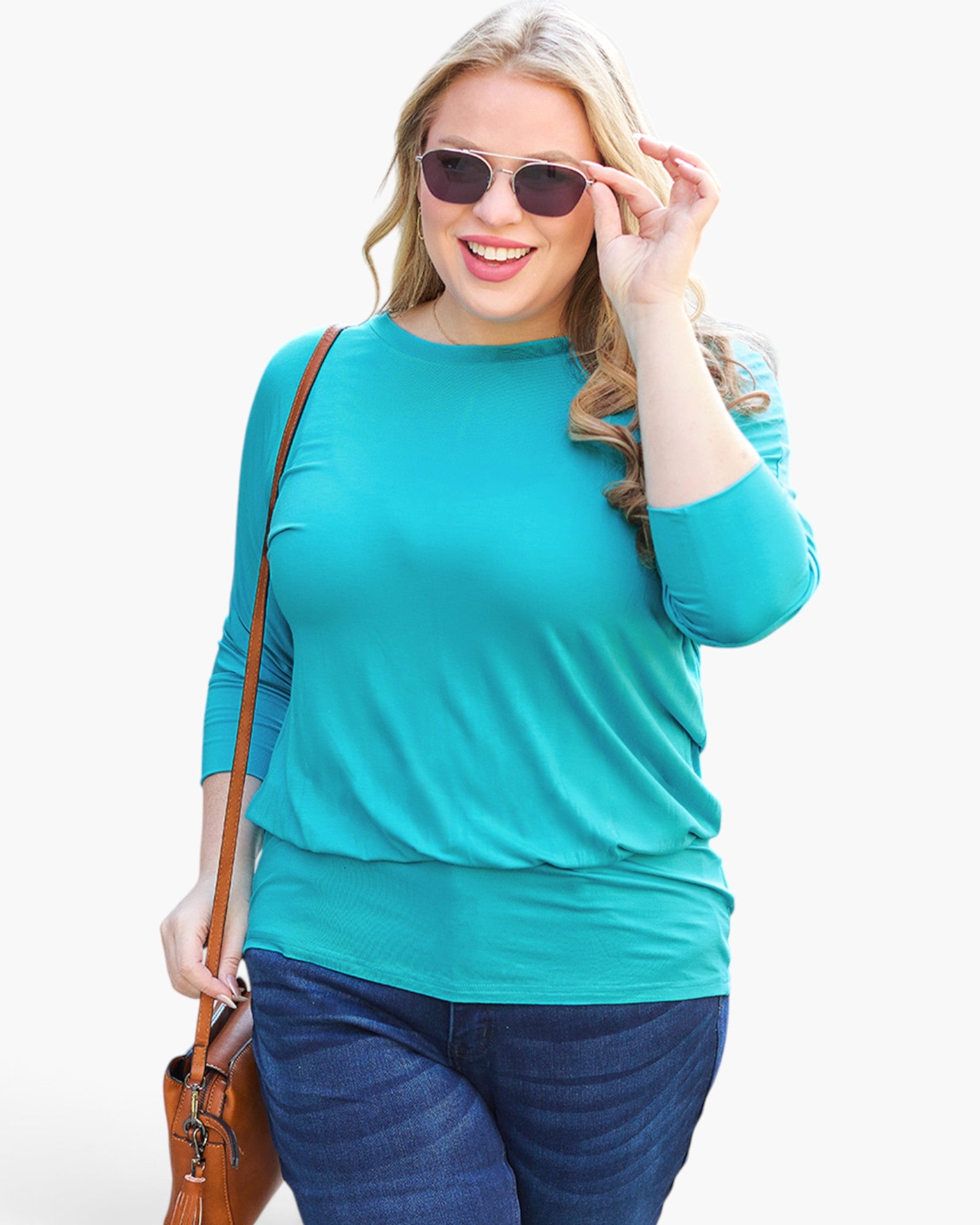 A woman modeling a bright turquoise three-quarter sleeve top with dark blue jeans, sunglasses, and a brown shoulder bag against a plain white background.