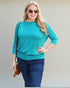 Woman wearing a turquoise top and blue jeans with sunglasses, standing against a plain background.