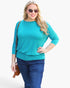 A woman modeling a turquoise three-quarter sleeve top with blue jeans, sunglasses, and a brown crossbody bag against a plain white background.