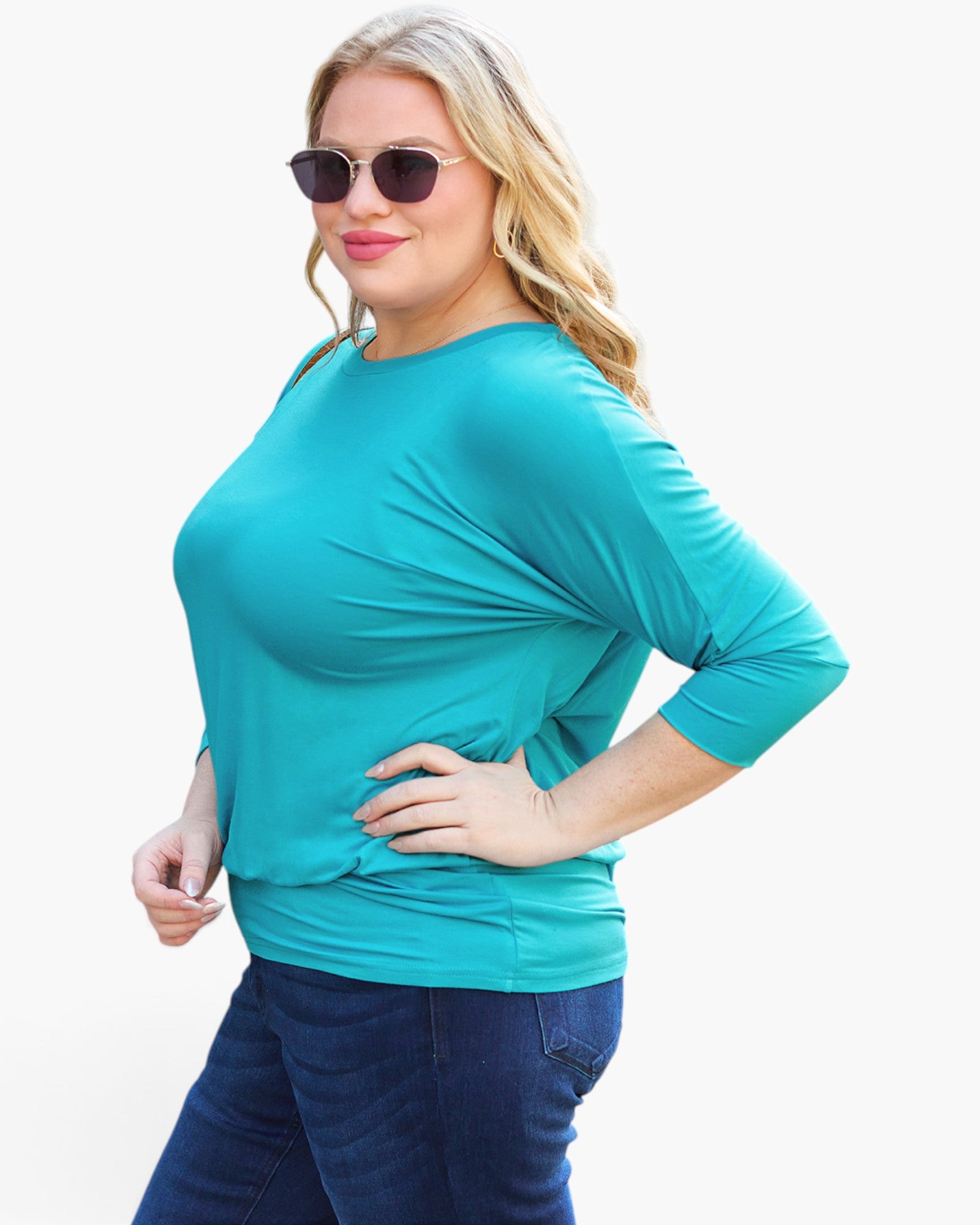 A woman modeling a bright turquoise three-quarter sleeve top with a boat neckline, paired with dark blue jeans and sunglasses, against a plain white background.