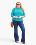 A woman modeling a turquoise three-quarter sleeve top and dark blue flared jeans, accessorized with a brown crossbody bag and sunglasses, against a plain white studio background.