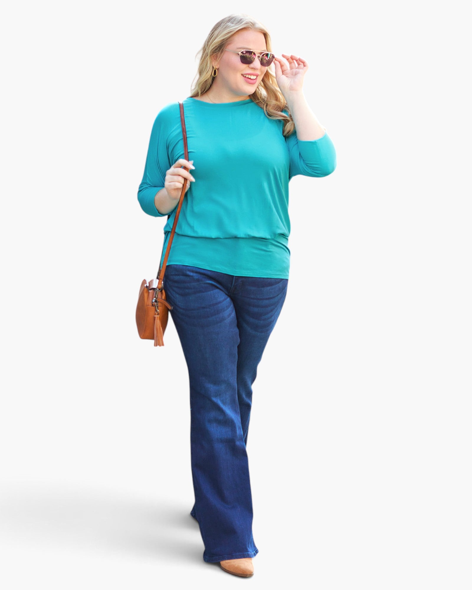 A woman modeling a bright turquoise long-sleeve top and dark blue jeans, carrying a brown leather crossbody bag against a plain white studio background.