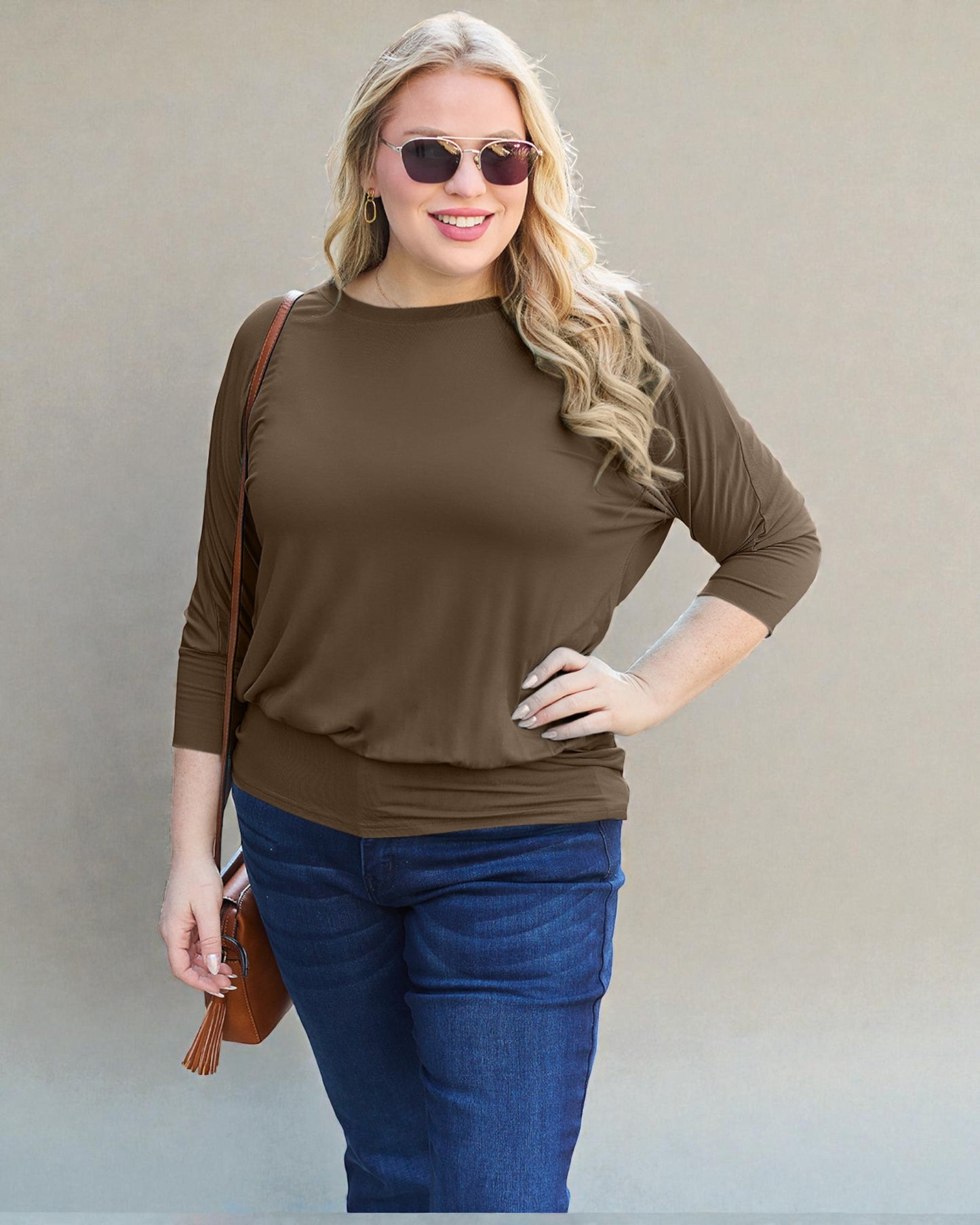 Woman wearing a brown top and blue jeans with sunglasses on a plain background