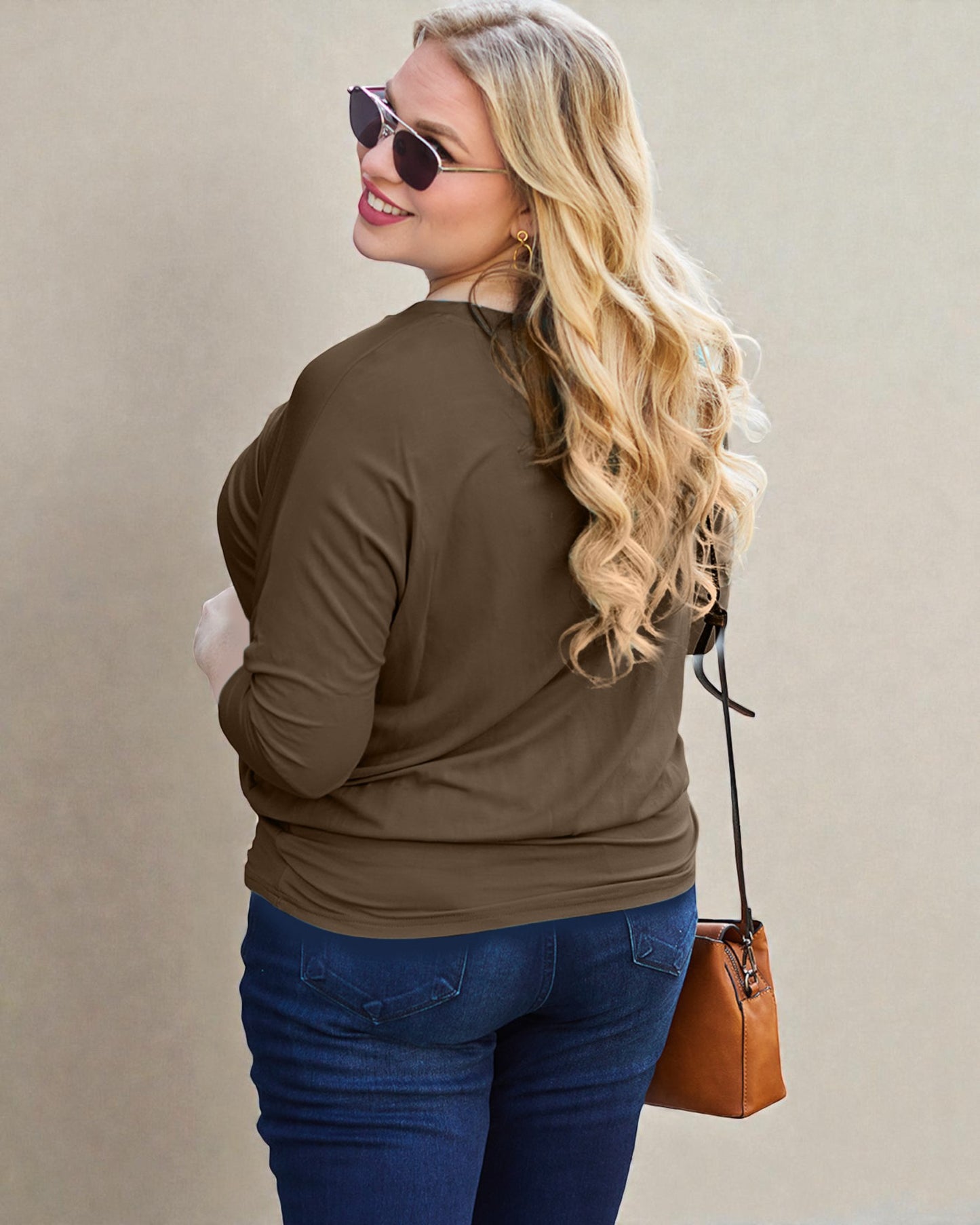 Woman wearing a brown long-sleeve top and blue jeans, holding a brown handbag against a beige background.