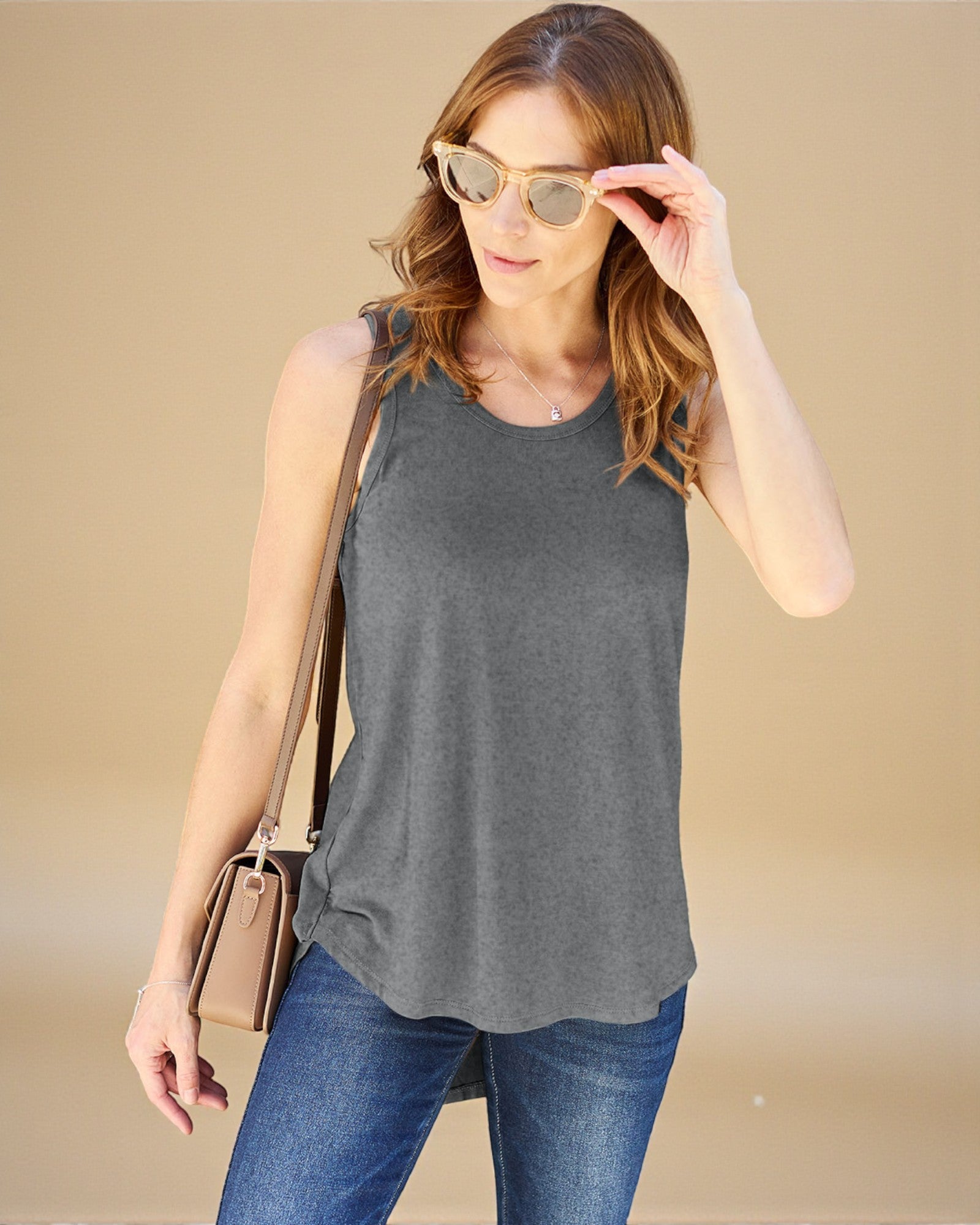 Woman wearing a gray sleeveless top and blue jeans, holding sunglasses and a beige handbag on a beige background.