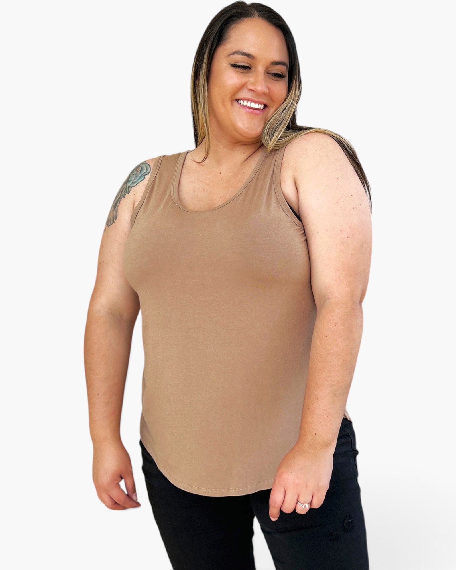 A smiling woman wearing a fitted scoop-neck tank top in a warm tan/beige color, paired with black pants, against a plain light gray studio background.