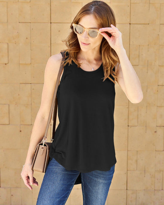 Woman wearing a black sleeveless top and blue jeans, holding sunglasses and a beige bag against a beige wall.