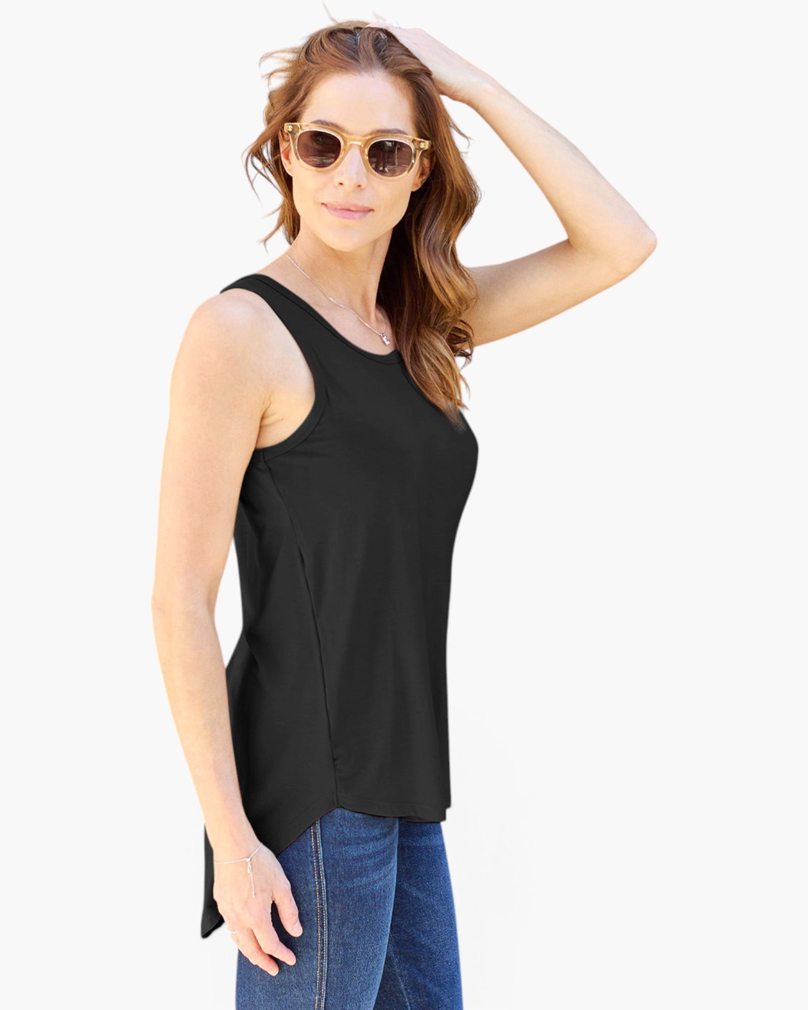 A woman modeling a black sleeveless scoop-neck tank top with a high-low hem, paired with blue jeans and round sunglasses, against a plain white background.