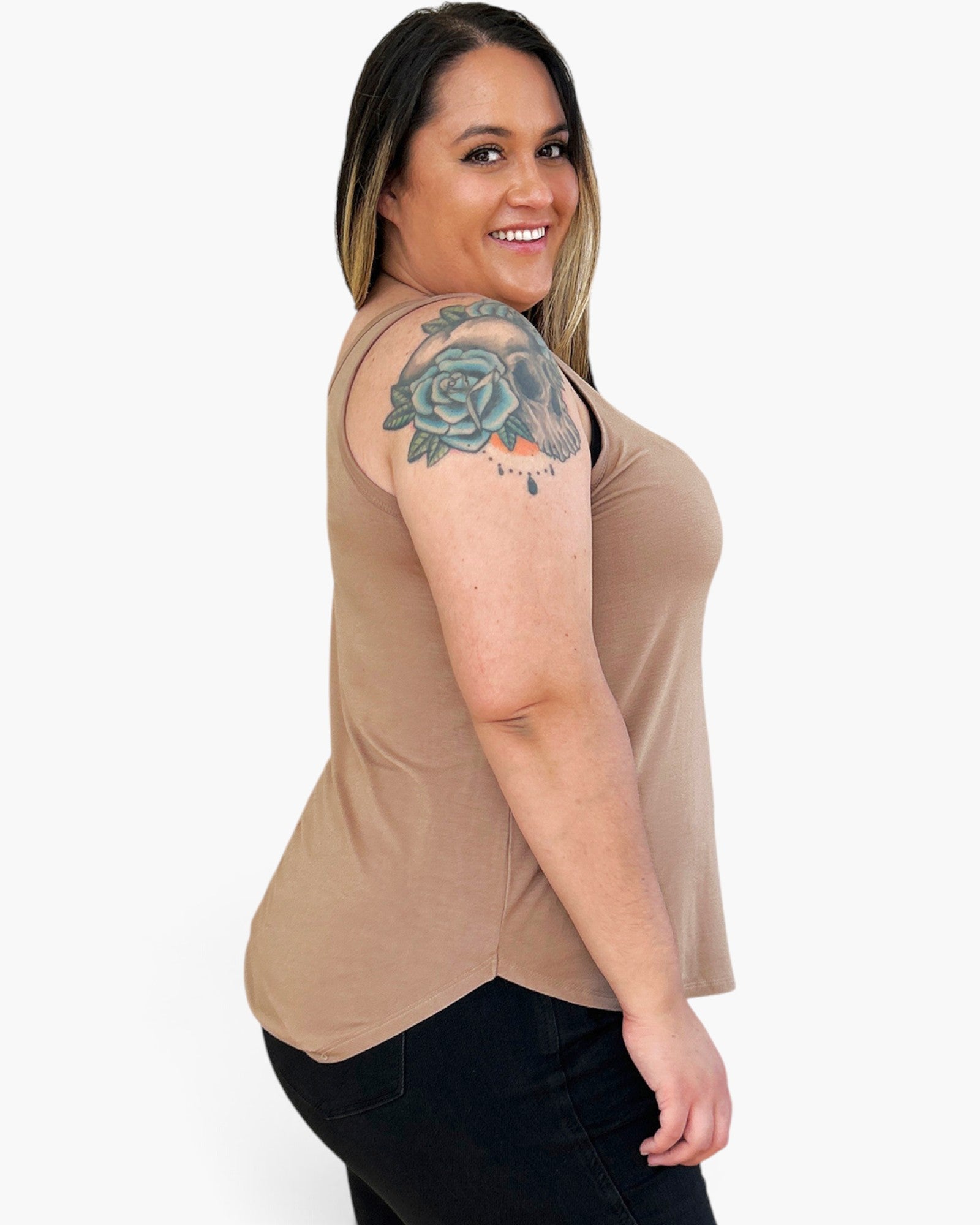 A woman with a floral tattoo on her upper arm is shown from the side/back wearing a sleeveless scoop-neck top in a warm tan/beige color, paired with black pants, against a plain light gray background.