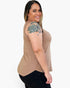 A woman with a floral tattoo on her upper arm is shown from the side/back wearing a sleeveless scoop-neck top in a warm tan/beige color, paired with black pants, against a plain light gray background.