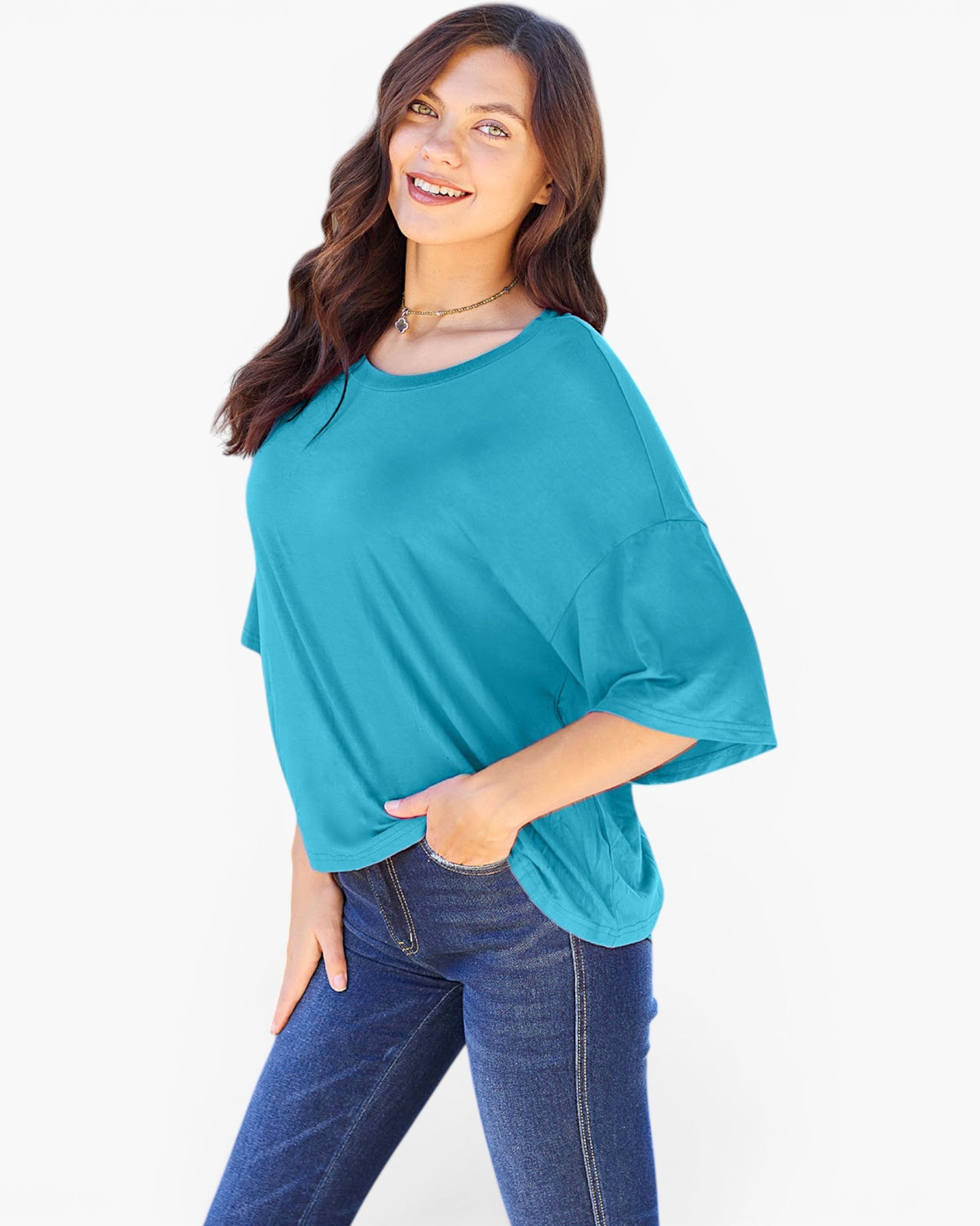 A woman modeling a teal blue short-sleeve top with a relaxed, oversized fit and side pockets, paired with dark blue jeans, against a plain white background.