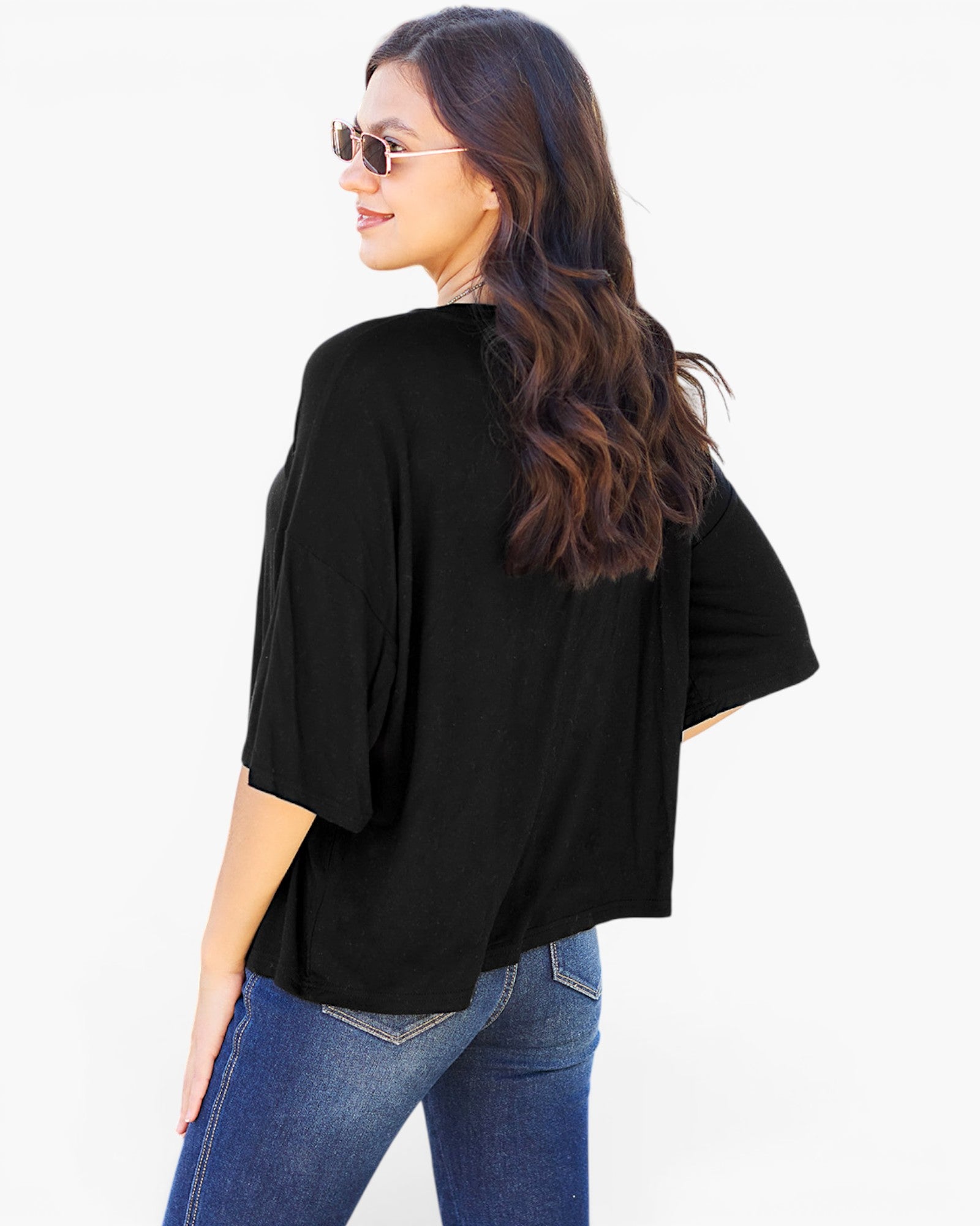 A woman with long wavy brown hair and sunglasses, wearing a loose black short-sleeve top and blue jeans, shown from a back-three-quarter angle against a plain white background.