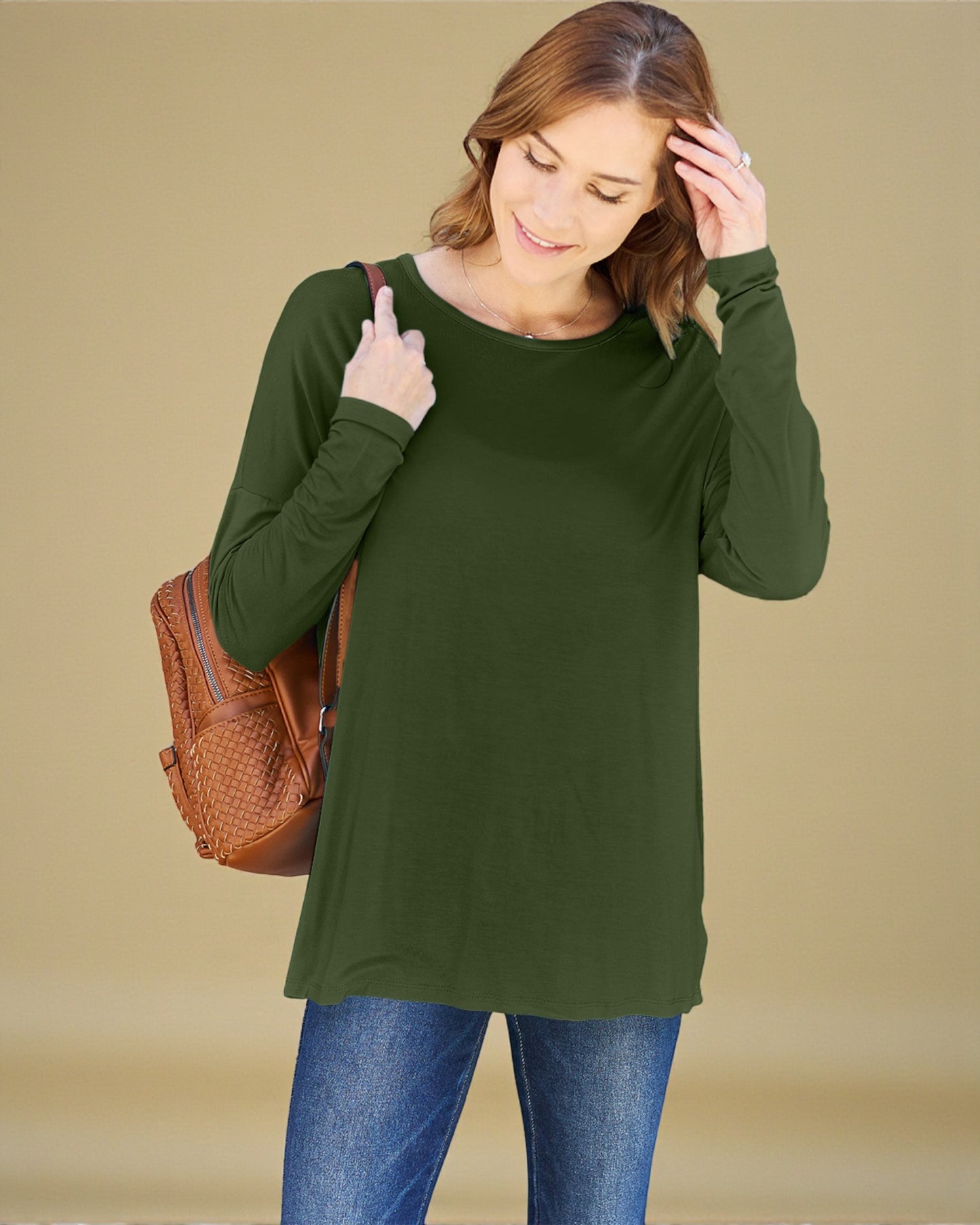 Woman wearing a green long-sleeve top and blue jeans, holding a brown backpack against a beige background.