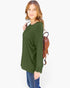 A woman wearing a long-sleeve olive green tunic top and blue jeans, carrying a brown woven backpack, photographed against a plain white studio background.