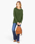 A woman modeling a long-sleeve olive green top, blue jeans, and brown shoes while holding a small brown woven leather backpack against a plain white background.