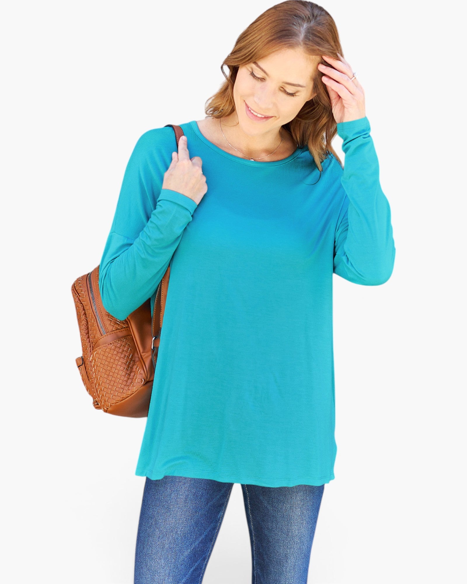 A woman wearing a bright turquoise long-sleeve tunic top and blue jeans, carrying a brown woven shoulder bag against a plain white background.