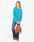 A woman modeling a bright turquoise long-sleeve top, blue jeans, and brown shoes, holding a small brown woven leather backpack against a plain white background.