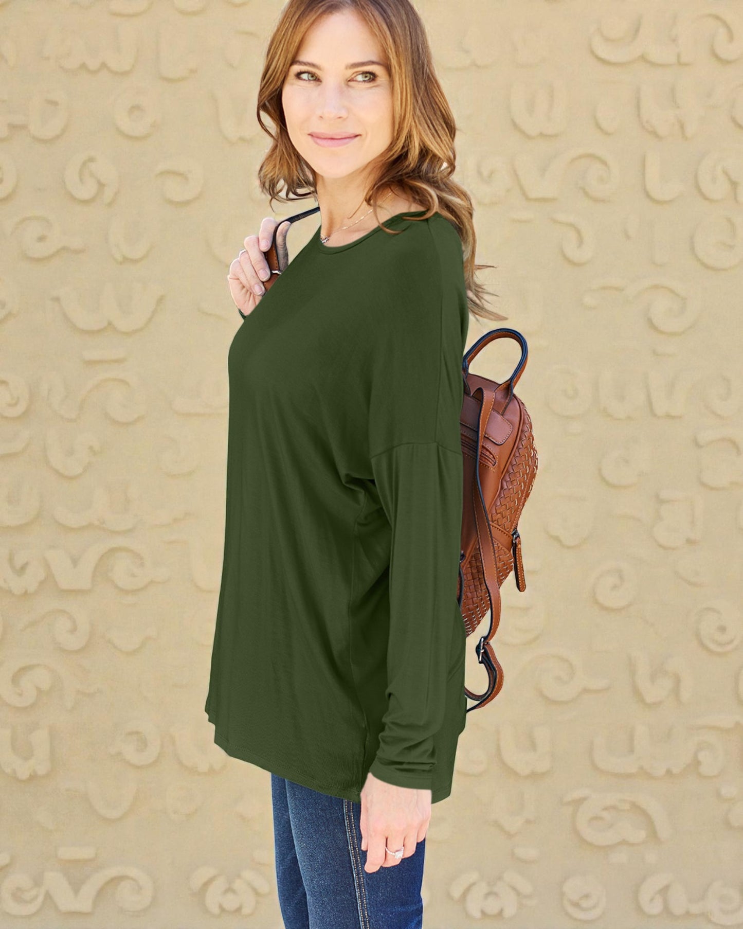 Woman wearing a green long-sleeve top with a brown backpack against a beige patterned wall.