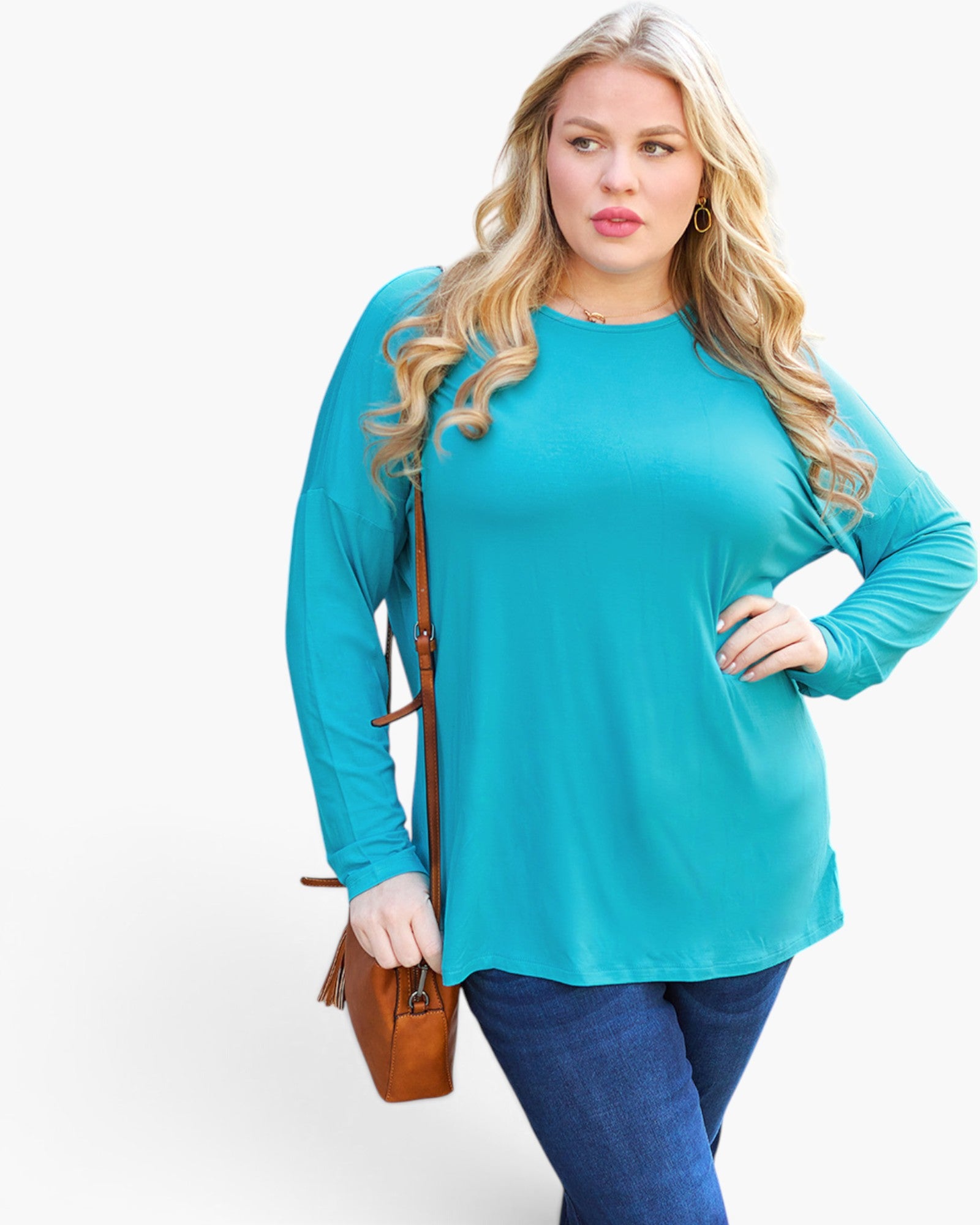 A woman modeling a bright turquoise long-sleeve top with blue jeans, holding a brown crossbody bag against a plain white studio background.
