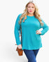 A woman modeling a bright turquoise long-sleeve top with blue jeans, holding a brown crossbody bag against a plain white studio background.