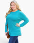 A woman modeling a bright turquoise long-sleeve top with blue jeans, holding a brown shoulder bag against a light gray studio background.
