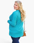 A woman with long blonde hair is shown from the back wearing a bright turquoise long-sleeve top and dark blue leggings, holding a brown crossbody purse against a plain white studio background.