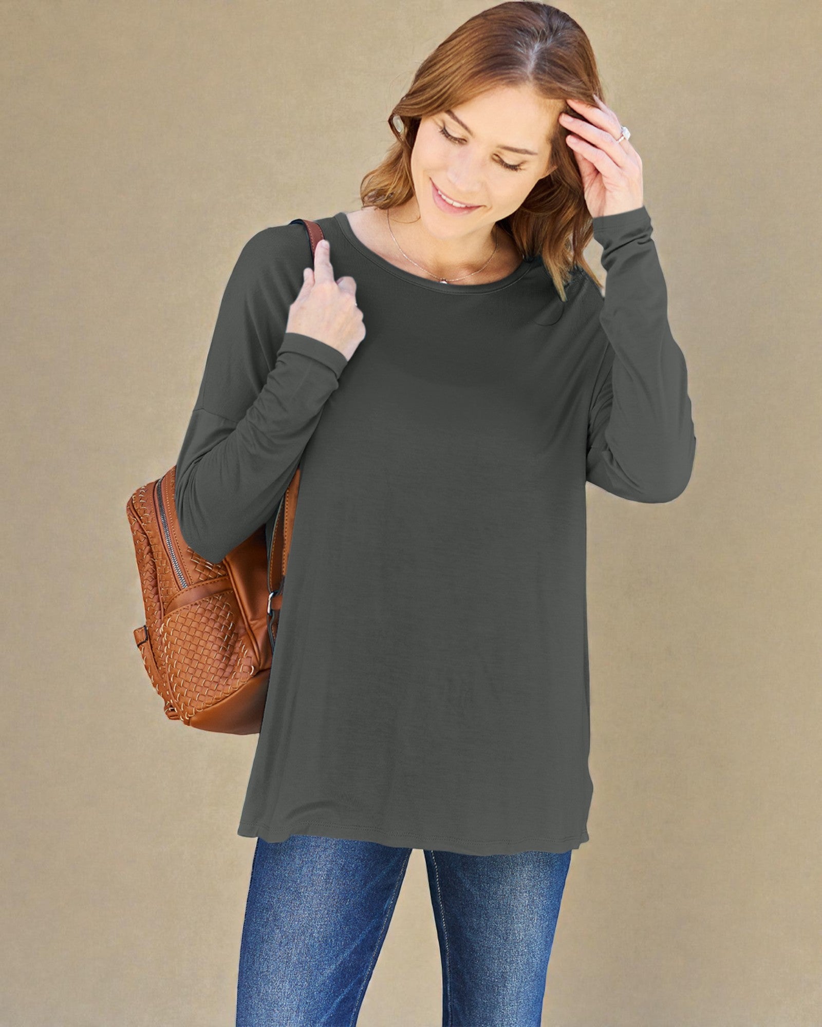 Woman wearing a dark gray long-sleeve top and blue jeans, holding a brown backpack against a beige background.