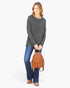 A woman modeling a dark gray long-sleeve top, blue jeans, and brown shoes while holding a brown woven leather mini backpack against a light gray studio background.