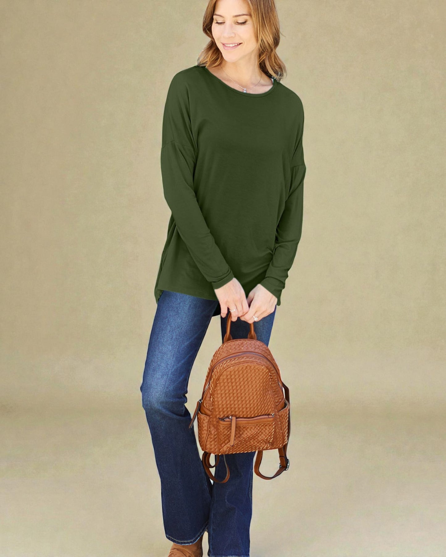 Woman holding a brown backpack wearing an olive green sweater and blue jeans on a beige background