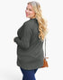 A woman modeling a dark gray long-sleeve tunic top with blue jeans, carrying a small brown crossbody bag, photographed against a plain light gray studio background.