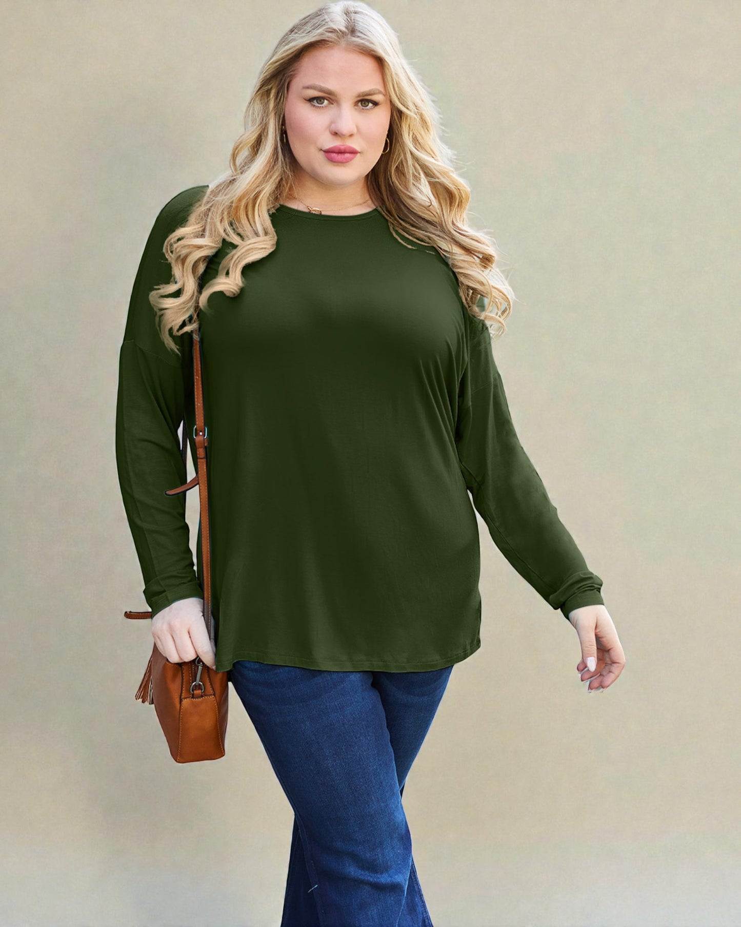 Woman wearing an olive green long-sleeve top and blue jeans, holding a brown leather bag against a beige background.