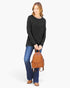 A woman modeling a black long-sleeve top, blue jeans, and brown shoes while holding a small brown woven leather backpack against a plain white background.