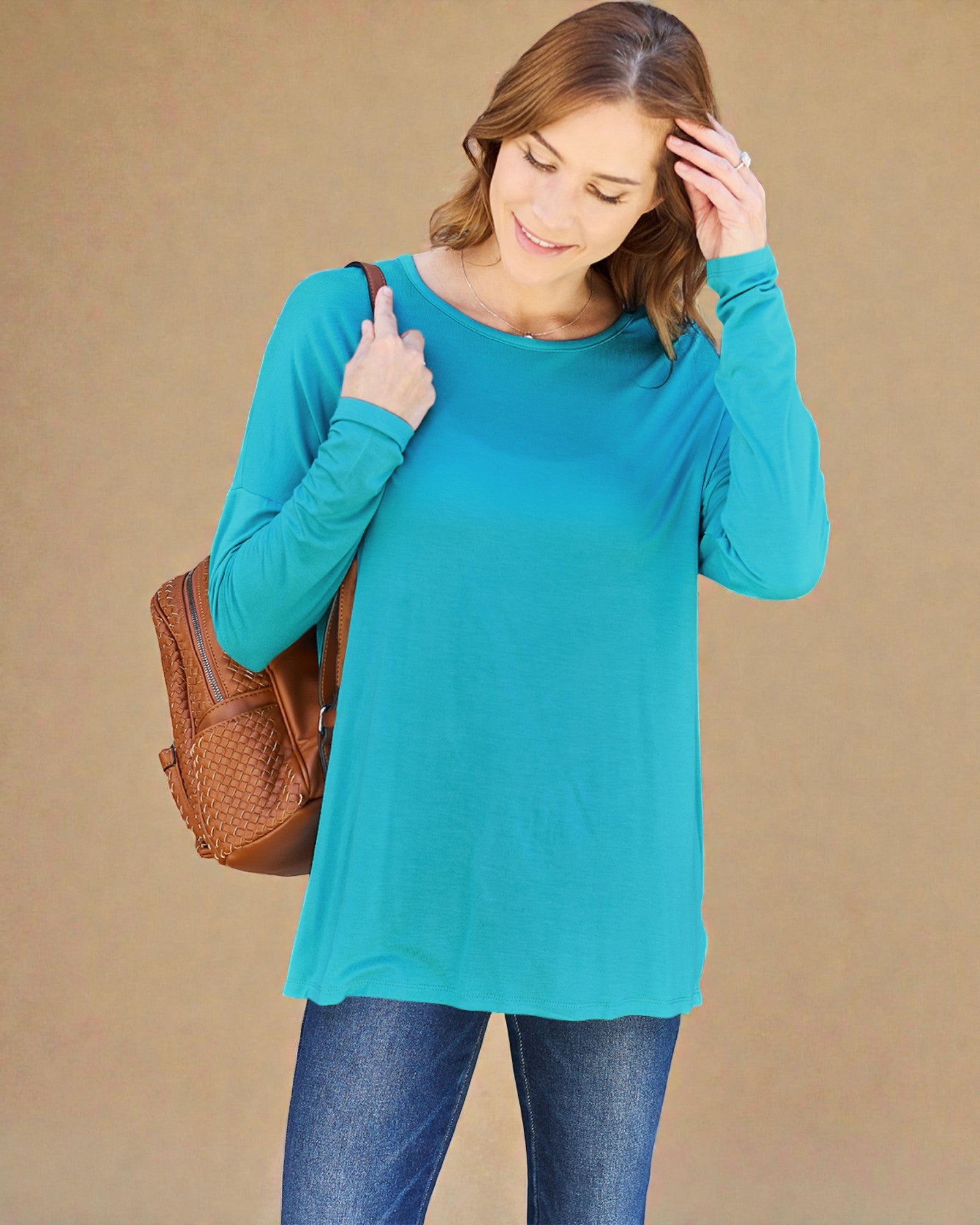 Woman wearing a teal long-sleeve top and blue jeans, holding a brown backpack against a beige background.