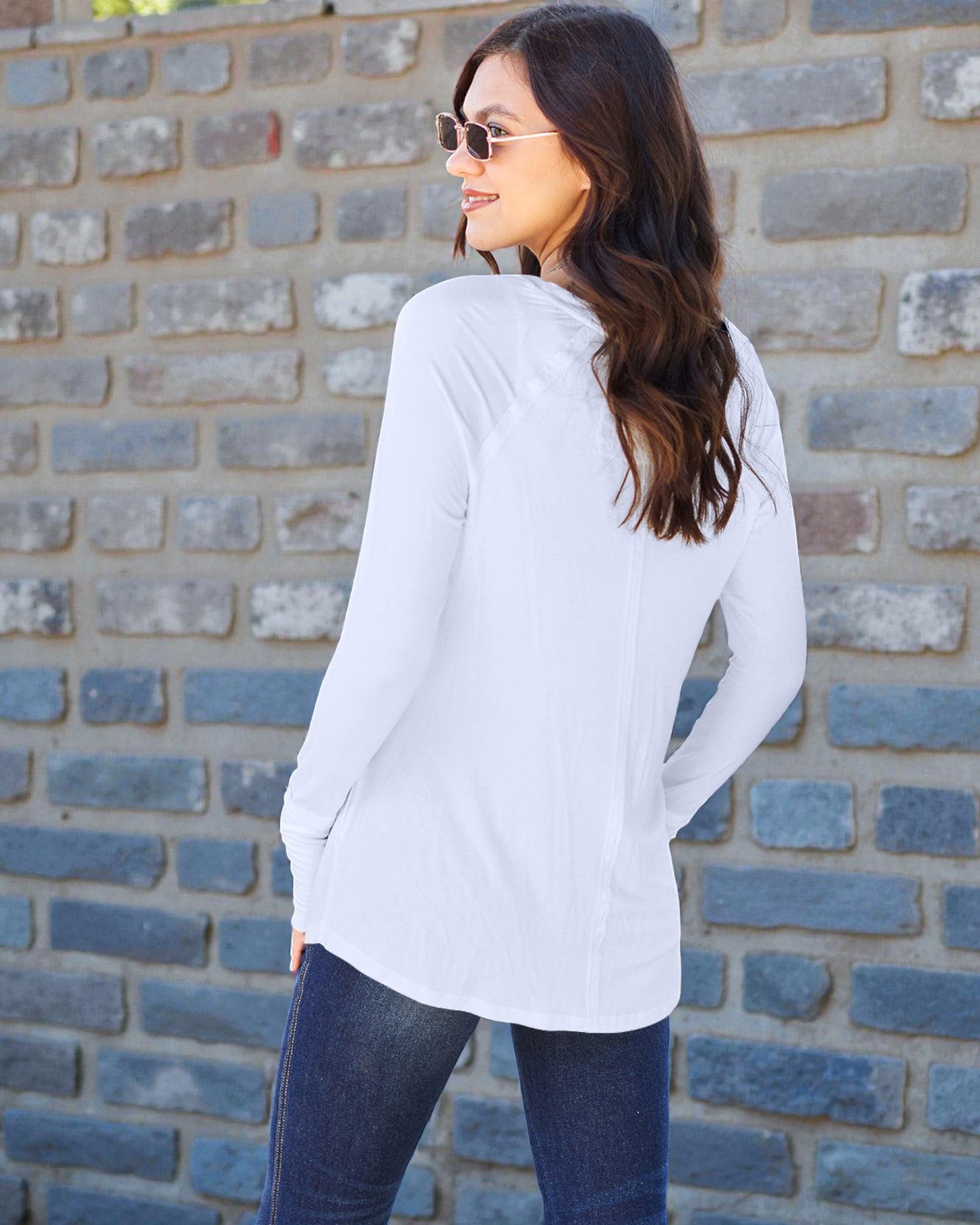 Woman wearing a white long-sleeve shirt and blue jeans against a brick wall.