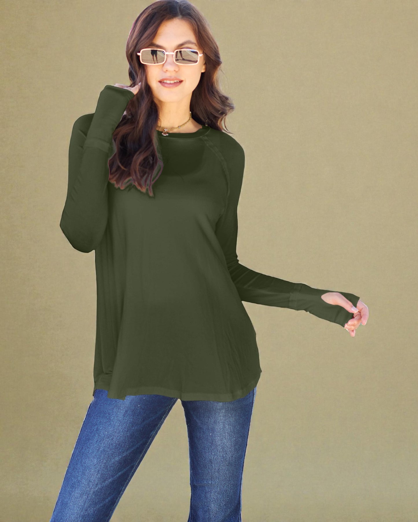 Woman wearing an olive green long-sleeve top and blue jeans against a beige background