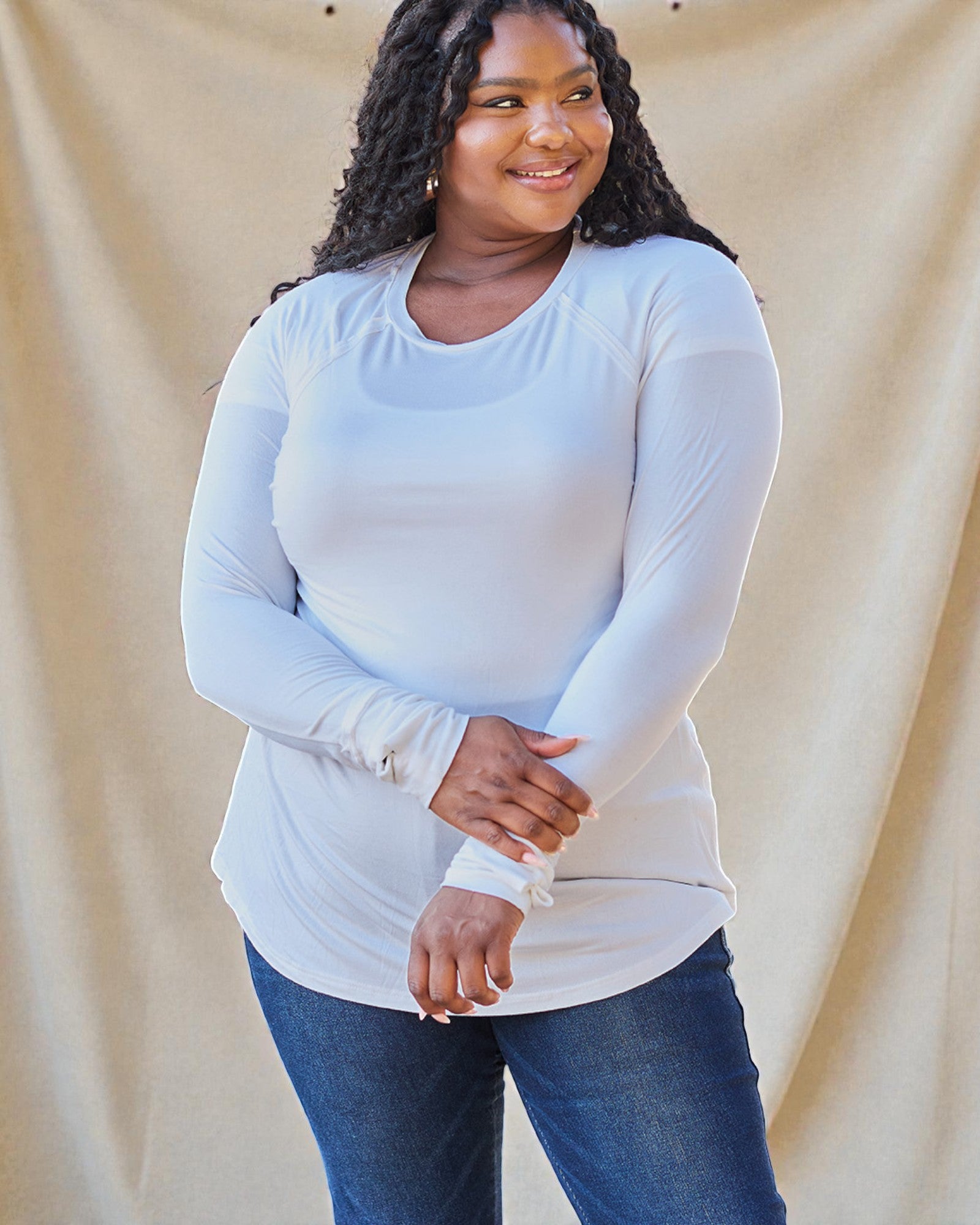 Woman wearing a white long-sleeve top and blue jeans against a beige background