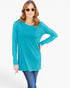 A woman modeling a bright turquoise long-sleeve tunic top with a single chest pocket, paired with blue jeans and gold-framed sunglasses, against a plain white studio background.