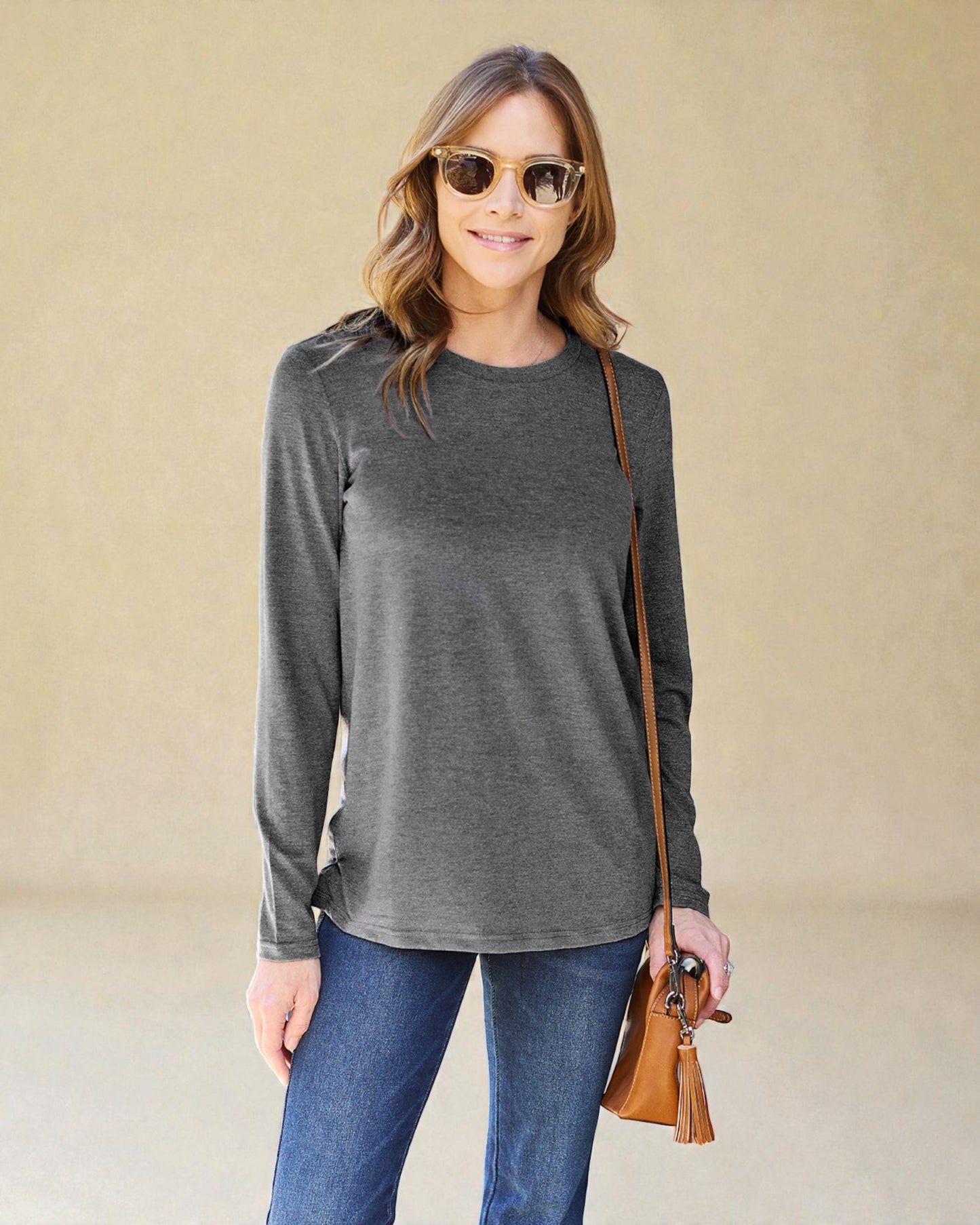 Woman wearing a gray long-sleeve top and blue jeans, holding a brown leather bag against a beige background.