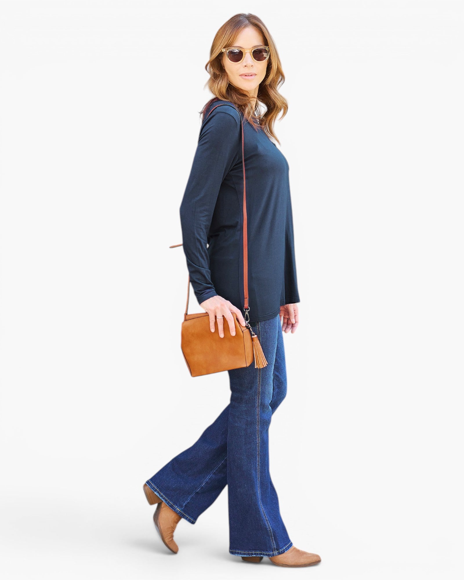 A woman modeling a navy blue long-sleeve tunic top with blue flared jeans, wearing a tan crossbody bag and sunglasses against a light gray background.