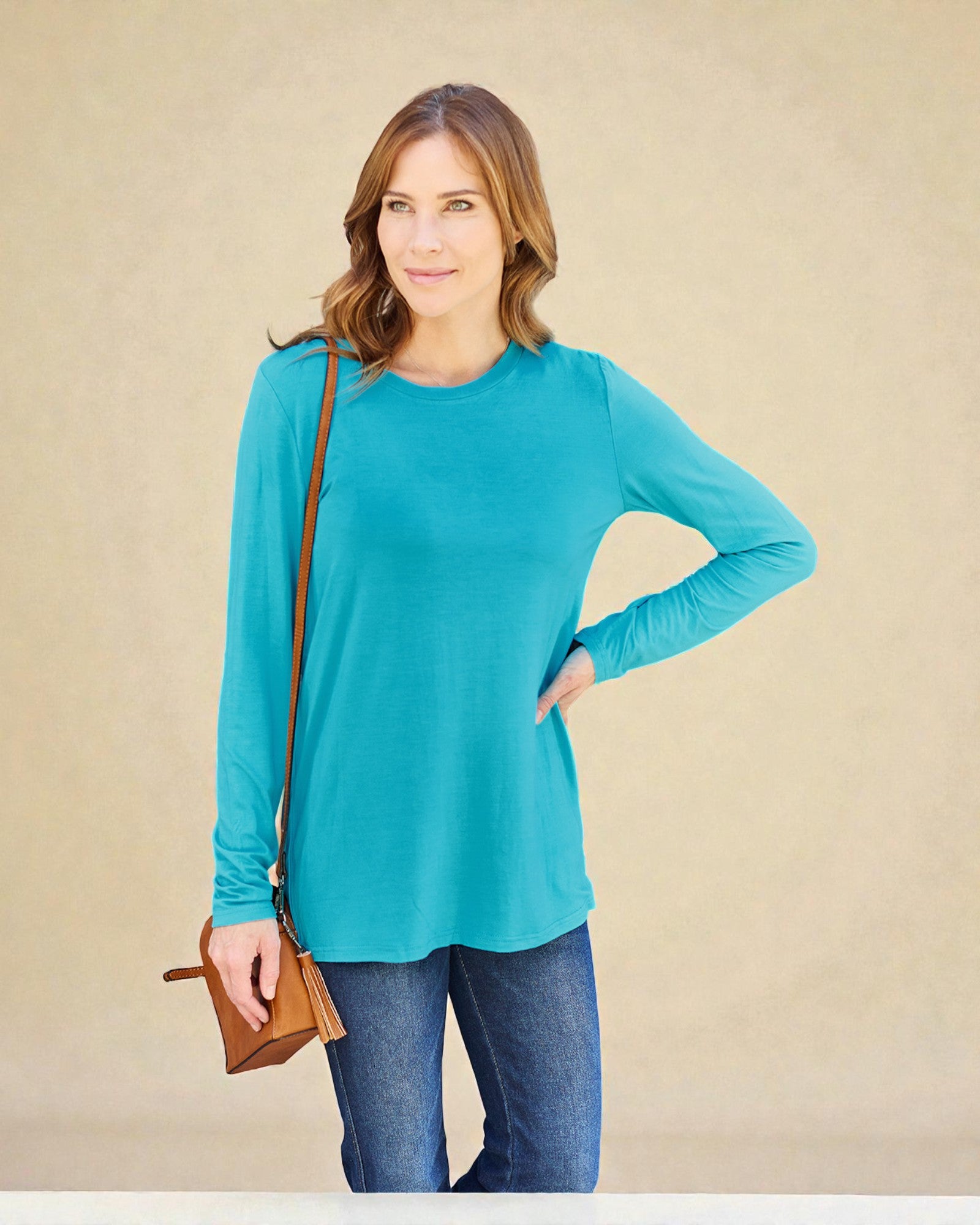 Woman wearing a teal long-sleeve top and blue jeans, holding a brown bag against a beige background